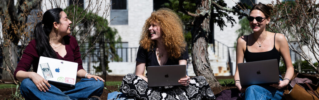 Three students sitting outdoors with laptops, smiling.